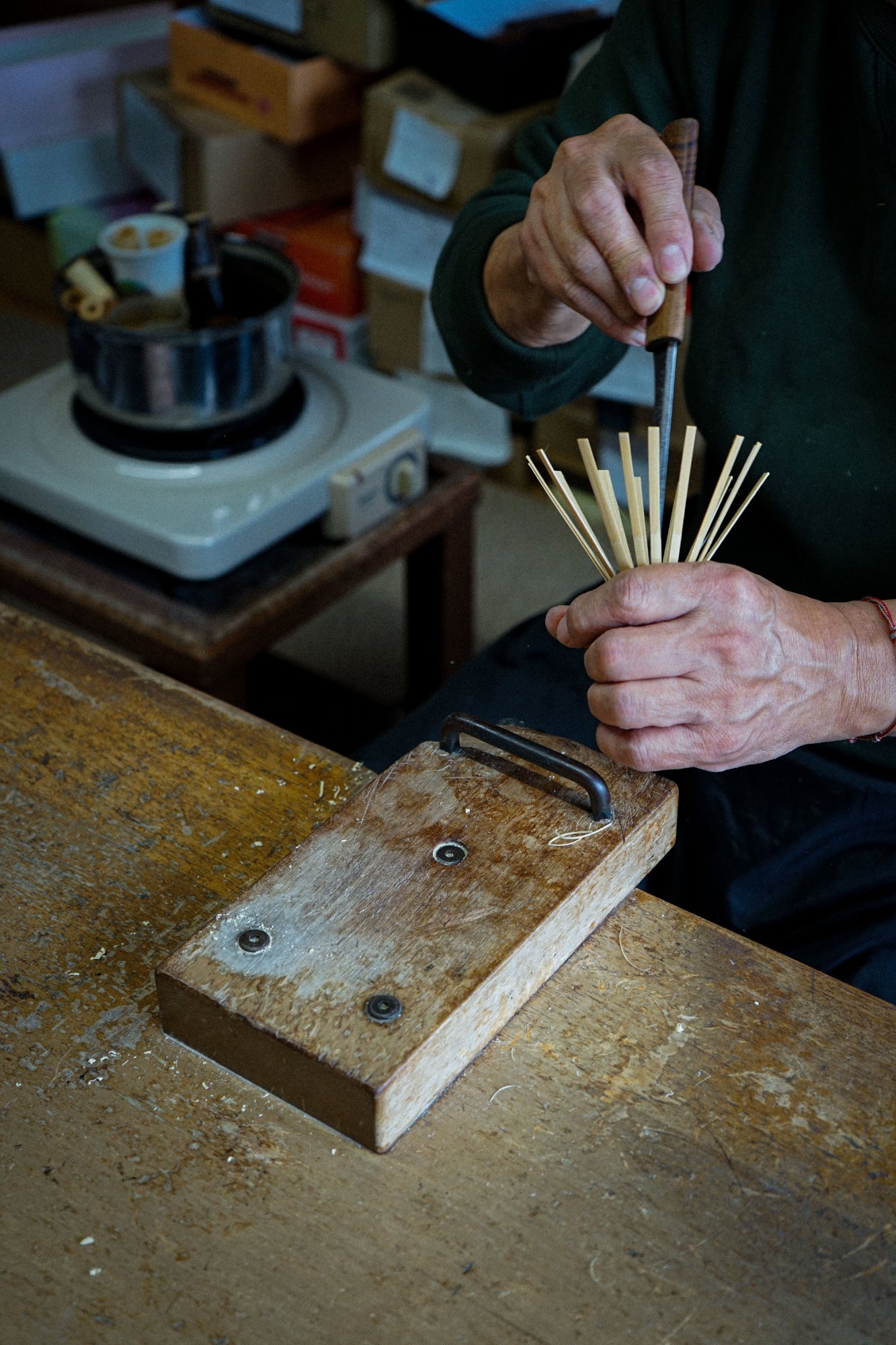 A close-up of a person's hands crafting small wooden tools or pins on a wooden workbench, with a portable burner and boxes in the background.
