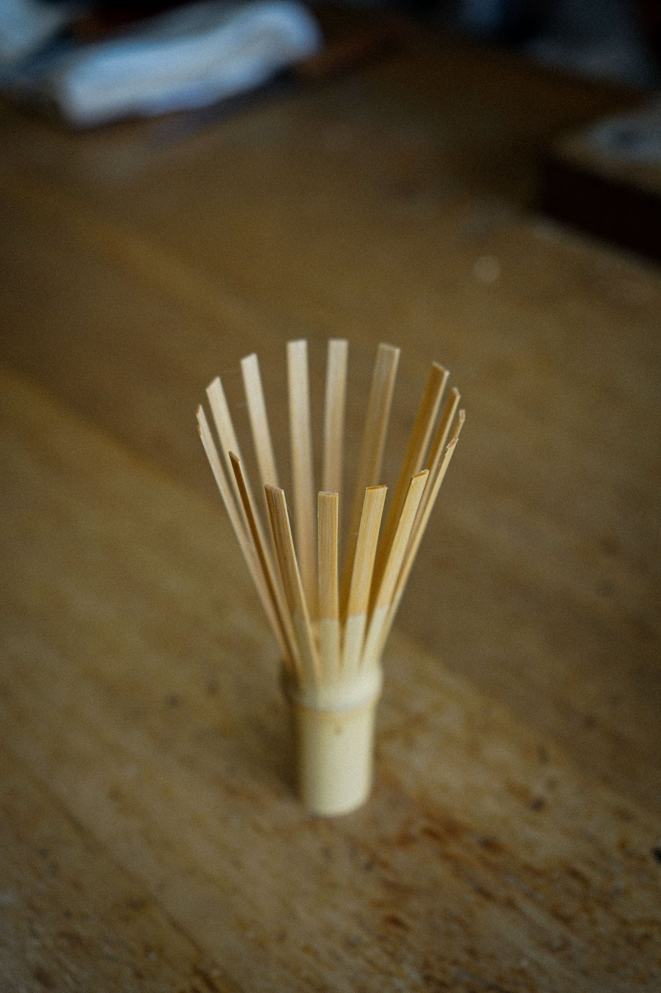 A small bamboo whisk with multiple thin bamboo sticks fanned out from a cylindrical handle, standing on a wooden tabletop.