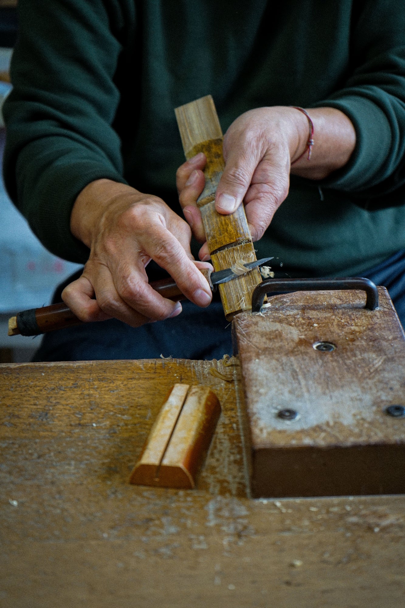 A close-up of a person's hands using a wooden carving tool on a piece of wood over a rustic wooden workbench.